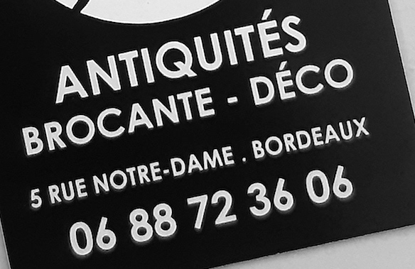 Un panneau noir et blanc affichant les mots 'ANTIQUITÉS BROCANTE - DÉCO' avec une adresse et un numéro de téléphone en bas.